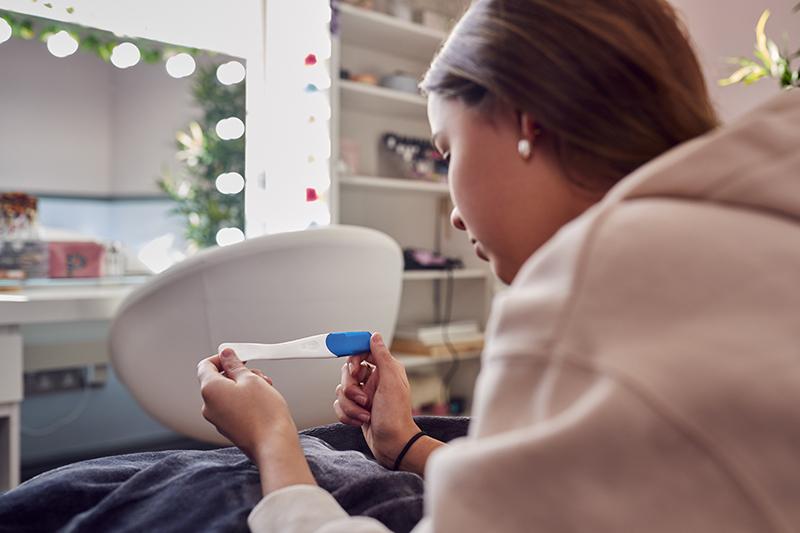 Close Up Of Teenage Girl Sitting On Bed At Home Looking At Positive Pregnancy Test